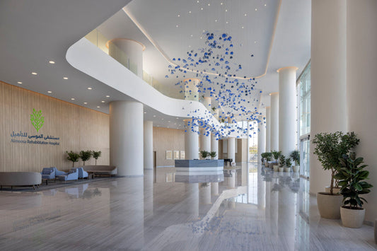 Modern hospital lobby interior at Almoosa Rehabilitation Hospital, featuring large white columns, floor-to-ceiling windows, elegant seating, and a decorative ceiling installation with blue hanging elements – ردهة مستشفى حديثة في مستشفى الموسى للتأهيل تحتوي على أعمدة بيضاء ونوافذ ممتدة من الأرض إلى السقف ومقاعد أنيقة وعناصر زينة معلقة باللون الأزرق 2