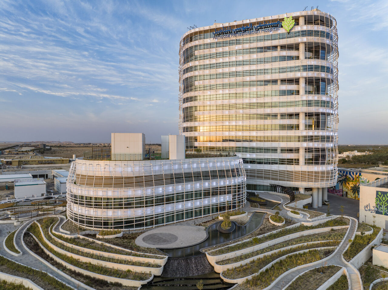 Exterior view of the Amoosa Rehabilitation Hospital featuring a modern architectural design with curved glass facades and tiered landscaping – healthcare facility in Saudi Arabia during sunset – منظر خارجي لمستشفى ألموسى للتأهيل بتصميم معماري عصري بواجهات زجاجية منحنية ومساحات خضراء متدرجة – منشأة رعاية صحية في المملكة العربية السعودية عند الغروب
