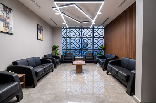 Modern reception lounge at Welaya in Riyadh featuring black leather sofas, a central wooden coffee table, wall art, indoor plants, and geometric mashrabiya-style windows. Contemporary lighting fixtures add elegance. صالة استقبال حديثة في مشروع ولاية بالرياض، تحتوي على كنبات جلدية سوداء، طاولة قهوة خشبية، إضاءة عصرية، نباتات داخلية، نوافذ بزخارف مشربية، وأرضية رخامية لامعة. 8