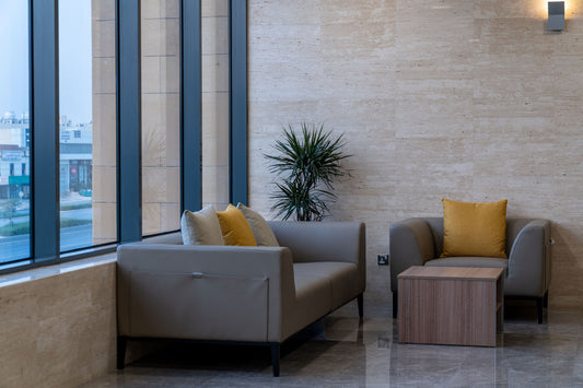 A quiet waiting area at Welaya in Riyadh featuring a modern grey sofa set with yellow and cream cushions, a wooden coffee table, and a green indoor plant. The natural stone wall and floor reflect the light from large windows overlooking the city. جلسة استقبال راقية في مشروع الولاية بالرياض تشمل كنبة حديثة، وسائد ملونة، طاولة قهوة خشبية، ونبات داخلي، تعكس الطابع المؤسسي الراقي والمريح في بيئة عمل رسمية. 3