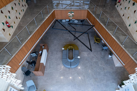 Top-down view of a modern lobby in Riyadh featuring a curved modular seating arrangement, wood-accented reception desk, and polished marble floors. The space includes contemporary office chairs, decorative lighting, and Saudi-style architectural elements. لقطة علوية لمنطقة استقبال في الرياض، بتصميم حديث يشمل جلسات دائرية، كراسي مكتبية، مكتب استقبال خشبي، أرضية رخامية، ولمسات هندسية سعودية تعكس طابع الفخامة المؤسسية. 2