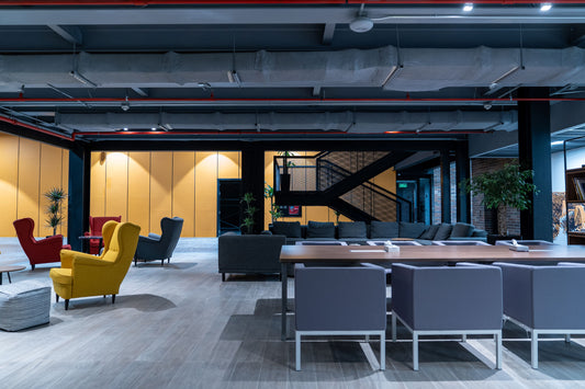 Lounge area at Misk Foundation in Riyadh with a mix of bold furniture including red, yellow, and grey lounge chairs, modular sofas, and meeting tables with grey armchairs. The space features industrial ceiling design, exposed red piping, and warm wooden flooring. Indoor greenery enhances the modern look. جمالية التصميم تعكس بيئة عمل متجددة وحيوية في قلب مدينة الرياض، توازن بين الراحة والإبداع داخل مساحة متعددة الاستخدامات. 5