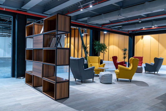 Modern lounge area at Misk Foundation in Riyadh showcasing vibrant armchairs in yellow, red, and grey, arranged around coffee tables and ottomans. The space features warm wood flooring, black metal open shelving, and industrial ceiling design with red piping. Indoor plants add greenery, while the lighting and color palette create a welcoming atmosphere. تصميم يجمع بين الأناقة والراحة يعكس بيئة عمل ملهمة في قلب الرياض. 4