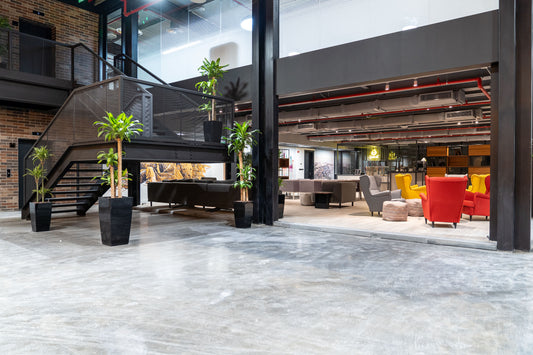 Industrial-style interior at Misk Foundation in Riyadh featuring exposed ceilings, modern black staircase, and vibrant lounge furniture in yellow, red, and grey. Potted green plants add warmth against concrete floors and brick walls. The open space blends collaborative zones and private meeting spots. التصميم يعكس بيئة عمل حديثة في الرياض تجمع بين العصرية والراحة، تعزز الإنتاجية والإبداع داخل مؤسسة مسك. 6
