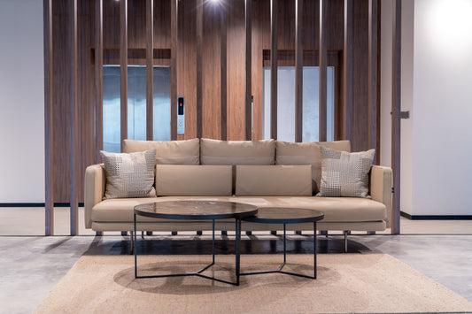 Modern lounge area at Azm Saudi Project, featuring a beige sofa with cushions, round coffee tables, and wooden wall panels, reflecting elegant interior design. Created by a Top Interior Designer in Riyadh and furnished by a Top Furniture Supplier in Saudi Arabia, the space combines comfort and sophistication. منطقة استراحة عصرية في مشروع عزم السعودية من عمل أفضل مصمم داخلي في السعودية مع أثاث من أفضل مورد أثاث في الرياض. 4