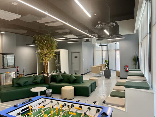 Recreational lounge at GDC, one of the PIF companies, featuring green modular sofas, foosball table, indoor tree, and acoustic ceiling panels – designed by Atelier 21 KSA, Top Interior Designer and Office Furniture Supplier in Saudi Arabia and Riyadh. صالة استراحة ترفيهية في مشروع GDC إحدى شركات صندوق الاستثمارات العامة، تضم أرائك مرنة وطاولة كرة قدم وزينة نباتية وسقفاً معالجاً صوتياً، من تصميم مفن الرسام، كأفضل مصمم داخلي ومورد أثاث ومصمم مكاتب في السعودية والرياض. 2