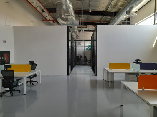 Workspace at Misk Foundation in Riyadh featuring clean white desks, black ergonomic chairs, and vibrant divider panels in orange, yellow, mint, and purple. The polished epoxy floor reflects natural light, while exposed ceilings and visible ductwork create an industrial-modern aesthetic. A central corridor with full-height glass walls leads to a conference room. تصميم حديث يعكس بيئة عمل إبداعية في مؤسسة مسك بالرياض. 10