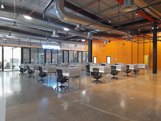 Open-plan workspace at Misk Foundation in Riyadh featuring industrial-style ceilings with exposed ducts, vibrant orange walls, and modern workstations equipped with ergonomic chairs. The polished concrete floor enhances the spacious layout. A glowing “INSPIRE” sign adds a motivational touch. تصميم مفتوح ومُلهم يعكس بيئة عمل مرنة وإبداعية، تعزز من التفاعل والإنتاجية في مقر مؤسسة مسك في الرياض. 8